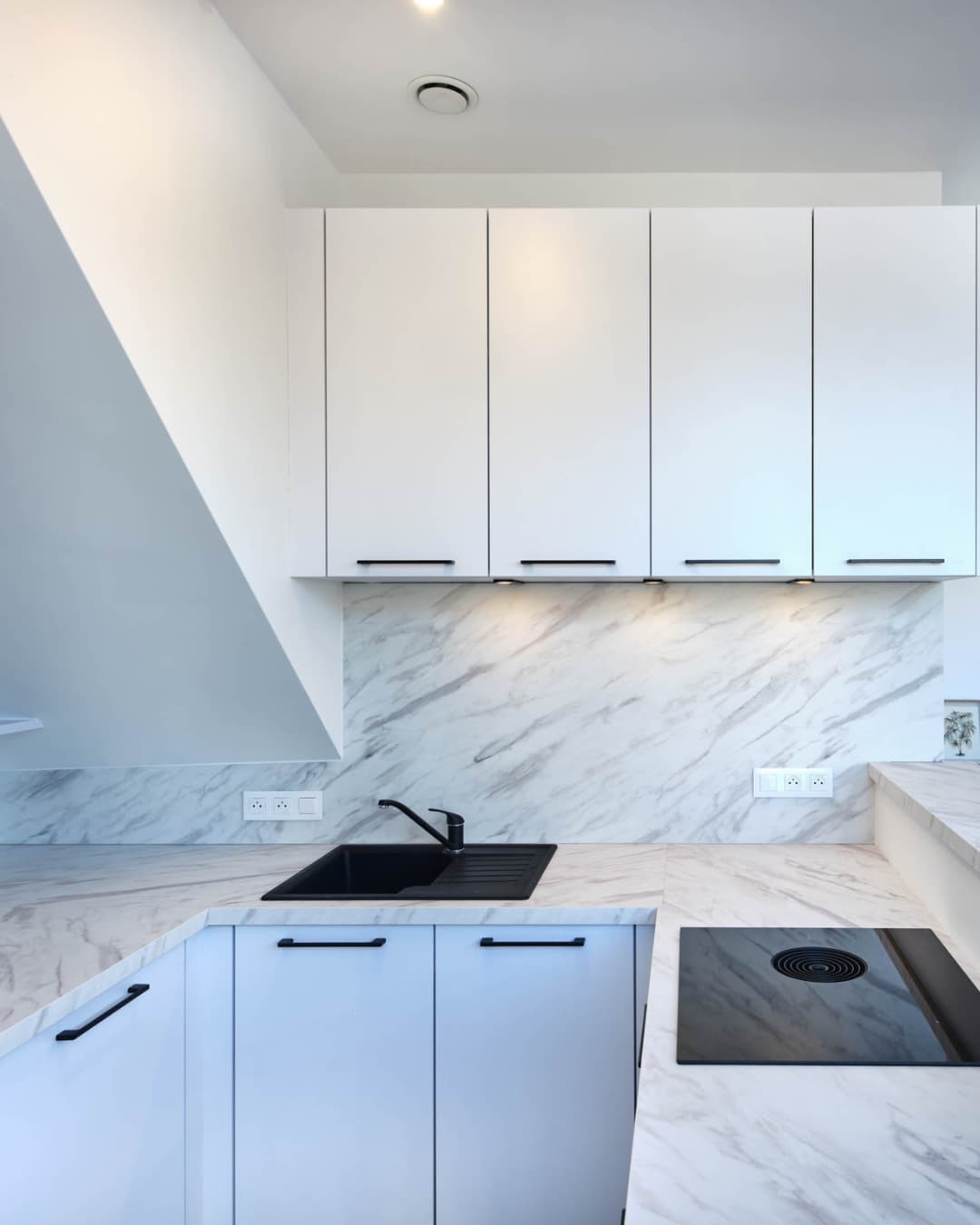 Petite cuisine en angle blanche avec rangements hauts sous-pente et plaque de cuisson à hotte intégrée pour l'optimisation de l'espace. IDÉO Cuisine