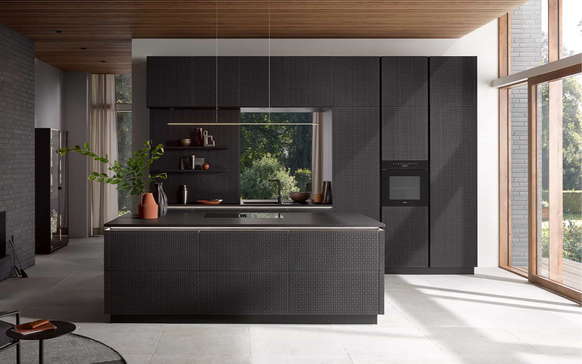 Cuisine design sur mesure noire avec façades texturées, îlot central avec hotte intégrée à la plaque de cuisson, four intégré en colonne et grandes baies vitrées. Design récompensé par le Best Of Design Award